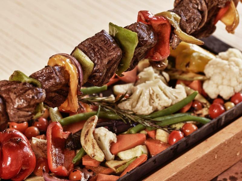 Gegrillte Fleischspieße mit buntem Gemüse auf Holzbrett, darunter Blumenkohl, grüne Bohnen und Petersilie.