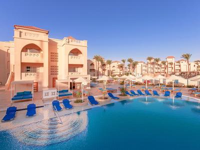 Hotel con grande piscina, lettini blu e edifici su più piani sotto cielo sereno.