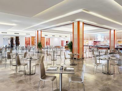 Moderner, heller Speisesaal mit vielen Tischen, Stühlen und warmen Holzsäulen und Beleuchtung.