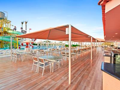 Area pranzo all'aperto con tavoli e sedie bianchi sotto una tenda rossa su una terrazza in legno.