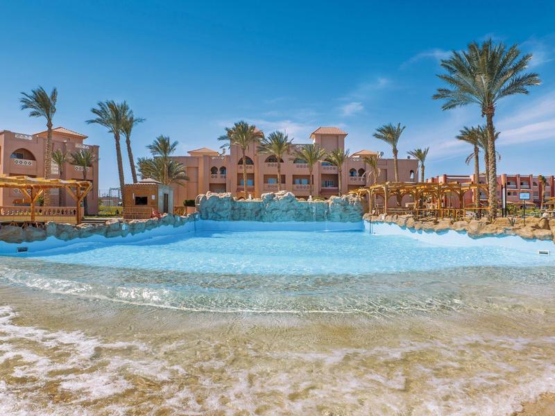 Großer Hotelpool mit Sandstrand, Palmen und mediterraner Architektur bei klarem Himmel.
