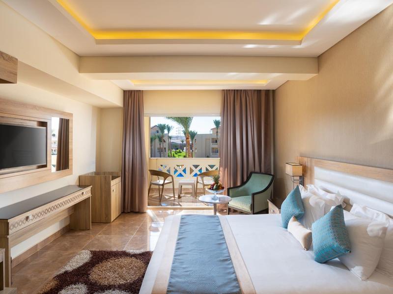 Helles Schlafzimmer mit großem Bett, blauen Kissen, TV, Schreibtisch und Balkon mit Pflanzen.