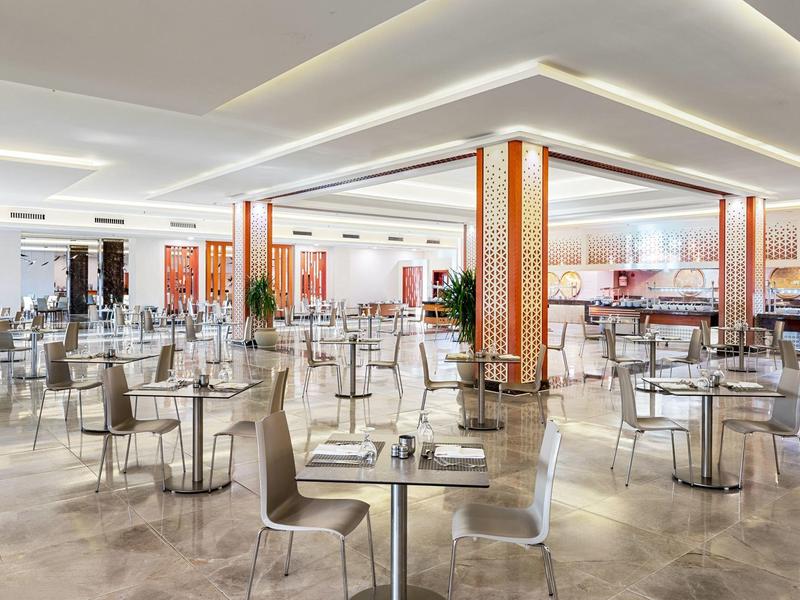 Moderner, heller Speisesaal mit vielen Tischen, Stühlen und warmen Holzsäulen und Beleuchtung.