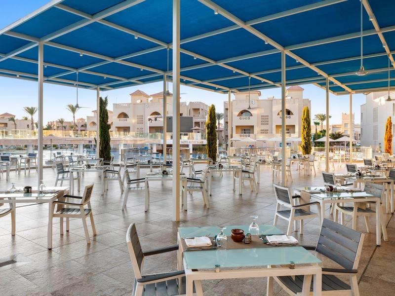 Große überdachte Terrasse mit vielen Metallstühlen und Tischen, Blick auf Wasser und Häuser.