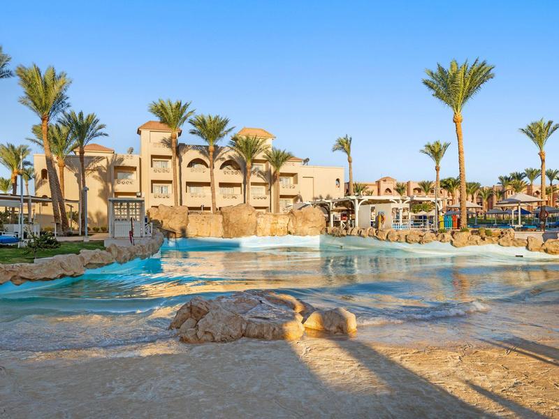 Großer Pool mit künstlichem Wasserfall, umgeben von Palmen und einem mehrstöckigen Hotelgebäude.
