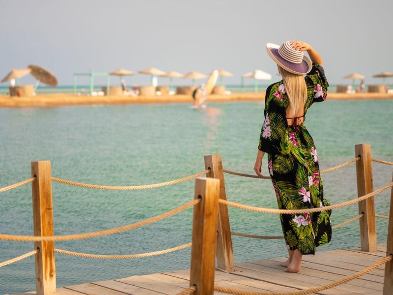 Femme en robe fleurie et chapeau regardant la mer depuis un ponton en bois.