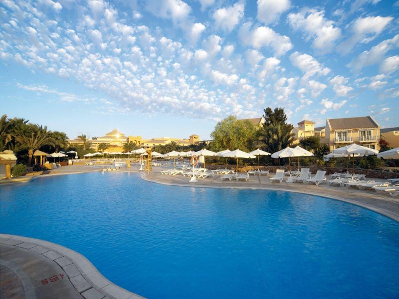 Piscine d'hôtel avec chaises longues et parasols sous un ciel bleu partiellement nuageux.