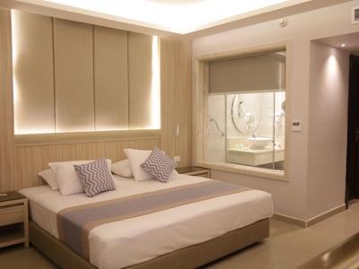 Modern hotel room with large double bed, pillows, and glass bathtub in the background