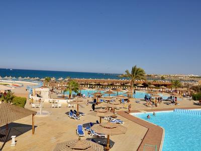 Blick auf einen Poolbereich mit Sonnenliegen, Sonnenschirmen und Meer im Hintergrund bei klarem Himmel.