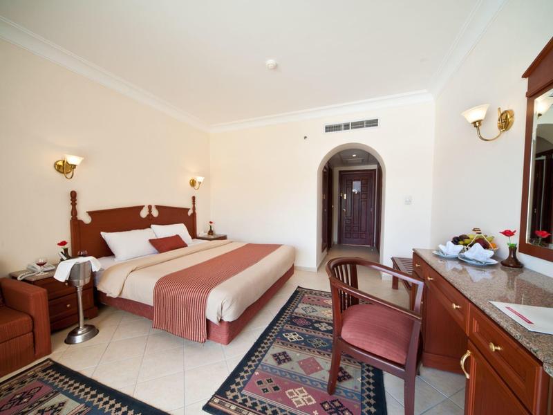 Helles Hotelzimmer mit Doppelbett, Teppichen, Stuhl, Sofa und Holzmöbeln mit großem Spiegel.