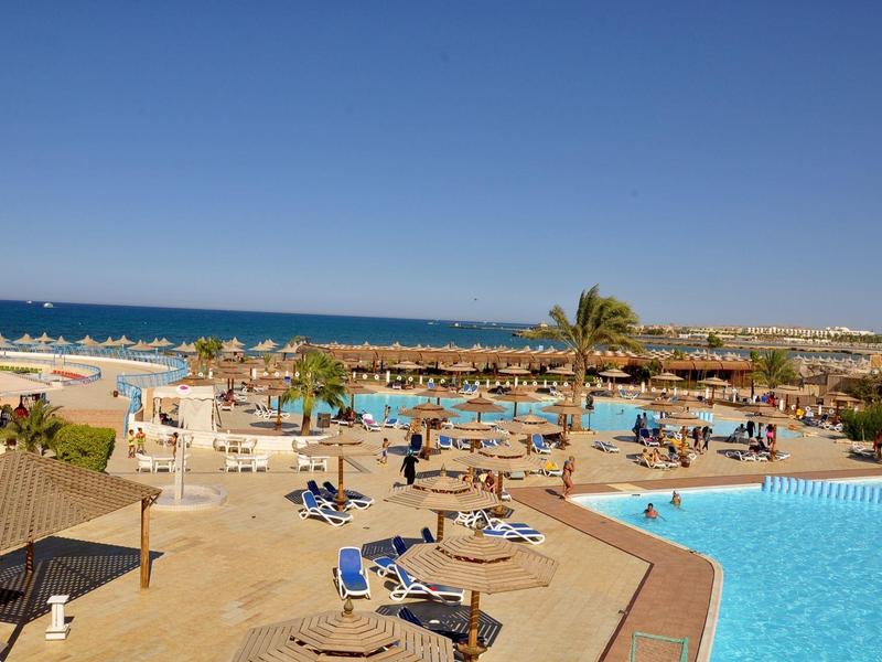 Blick auf einen Poolbereich mit Sonnenliegen, Sonnenschirmen und Meer im Hintergrund bei klarem Himmel.