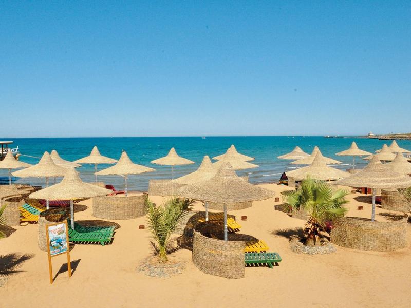 Strand mit vielen Strohhut-Sonnenschirmen, Liegen und Palmen vor ruhigem blauem Meer.