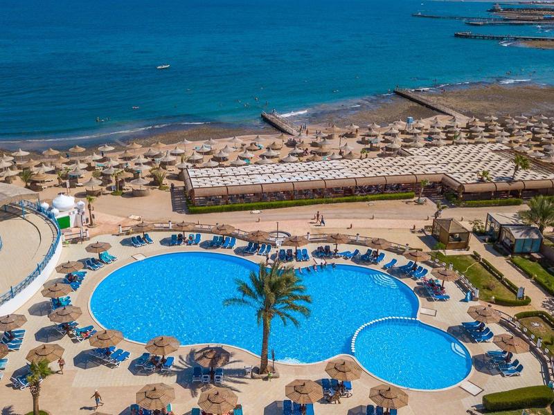 Großer Hotelpool mit Liegestühlen, Sonnenschirmen und Blick auf das Meer und die Küste.