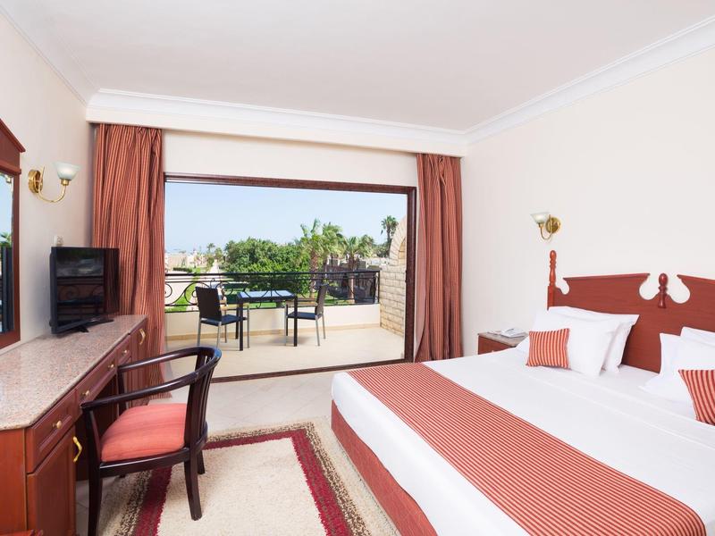 Hell eingerichtetes Hotelzimmer mit großem Bett, Schreibtisch, Stuhl und Balkon mit Gartenblick.