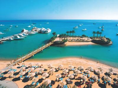 Playa con sombrillas y un muelle que conduce a una pequeña isla con palmeras y barcos en el mar.