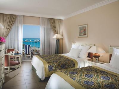 Hotel room with twin beds, elegant decor, and balcony overlooking the sea and outdoor seating.
