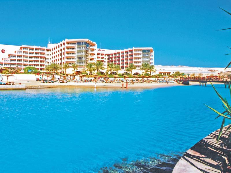 Large hotel building by a bright blue pool with palm trees and clear sky.