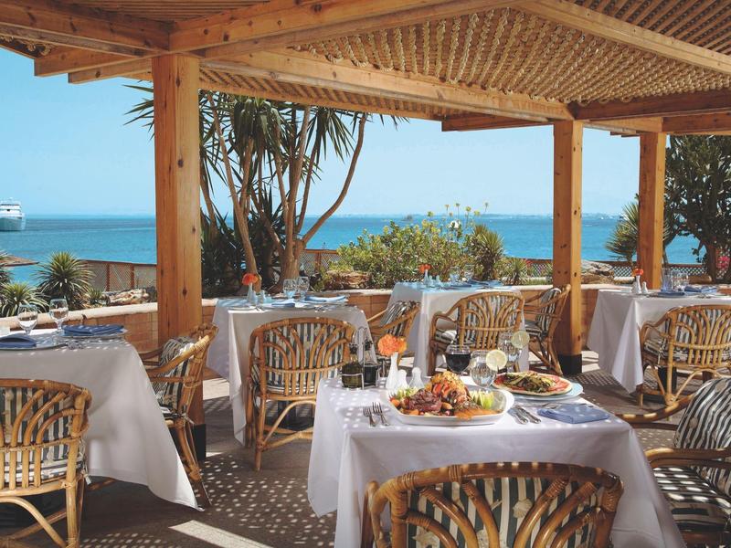 Outdoor dining area with tables, chairs, and sea view under a wooden pergola.
