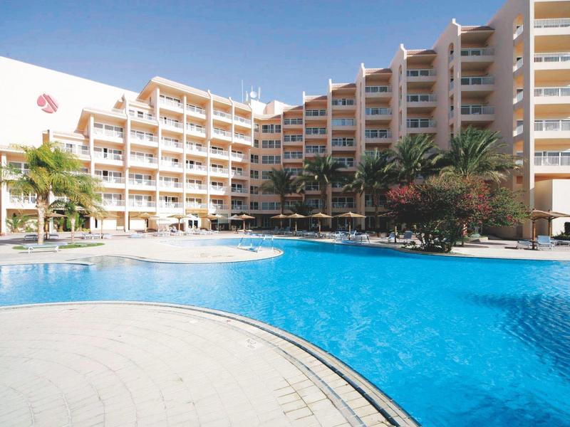 Large hotel building with multiple balconies beside a spacious outdoor swimming pool under clear blue sky.