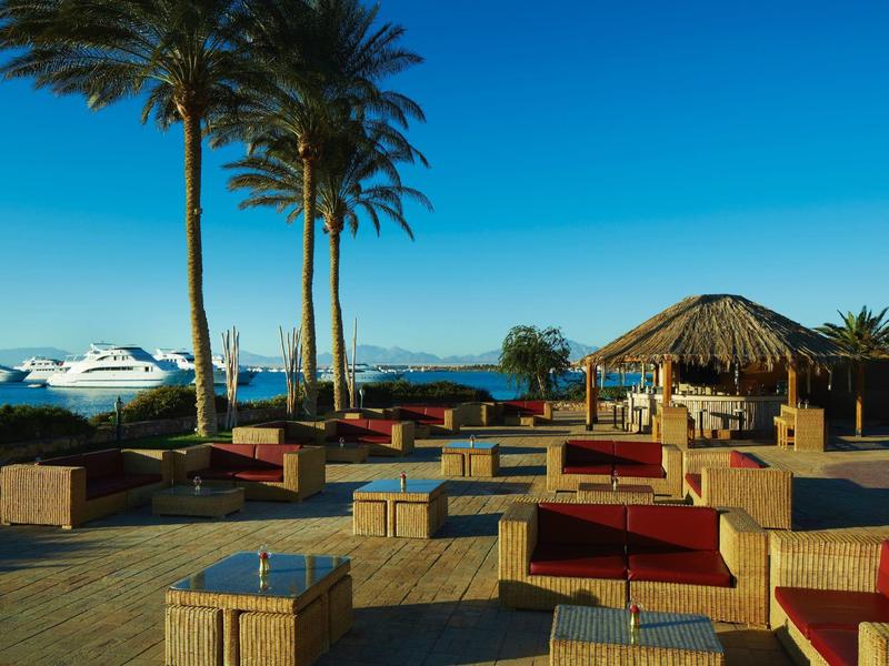 Outdoor lounge area with wicker seating, palm trees, and boats in the sea under clear sky.