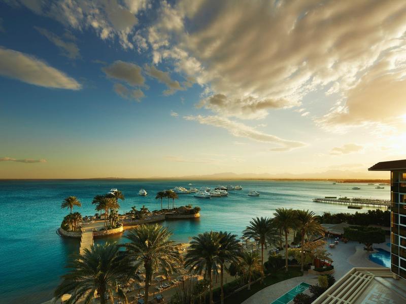 Ocean view with palm trees, boats, and a sunset sky over a resort.