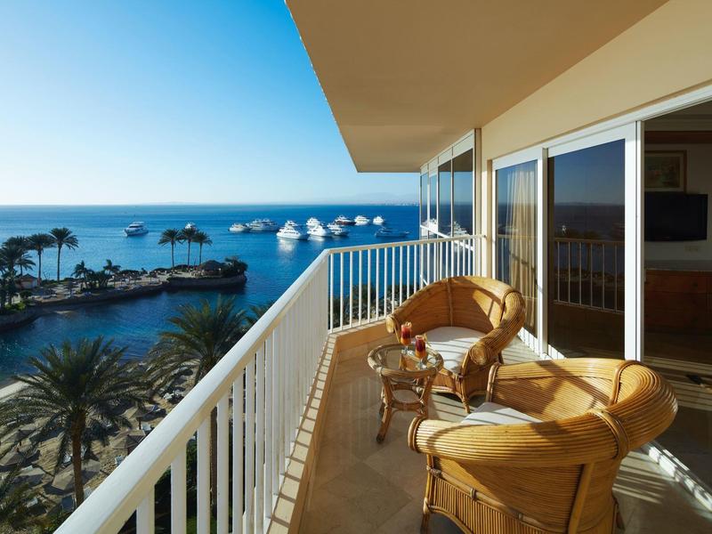 Balcony with chairs overlooking ocean, boats, and palm trees on a sunny day.