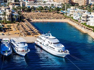 Luxury yachts docked at a harbor near a beach with sun umbrellas and white buildings.
