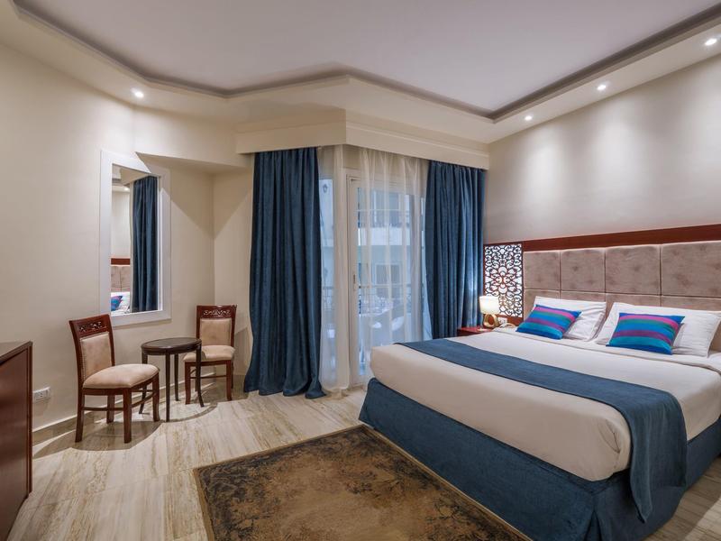 Schlafzimmer mit großem Bett, blauen Vorhängen, Holzstuhl, Tisch und hellen Wänden.