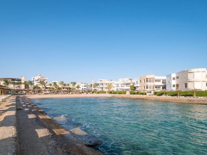 Klares Meer mit Promenade, weißen Gebäuden und blauem Himmel im sonnigen Küstenort.