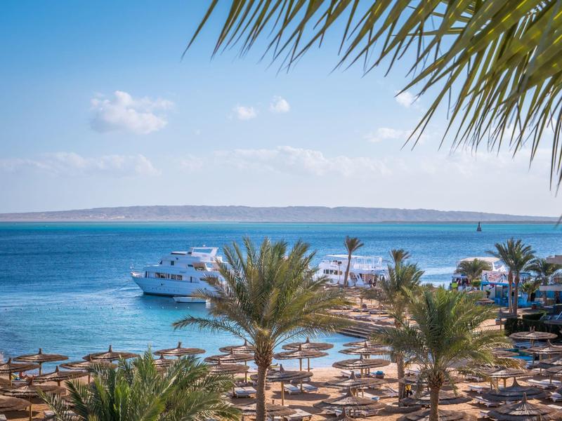 Meerblick mit Yachten, Palmen und Sonnenschirmen unter klarem Himmel am Strand.
