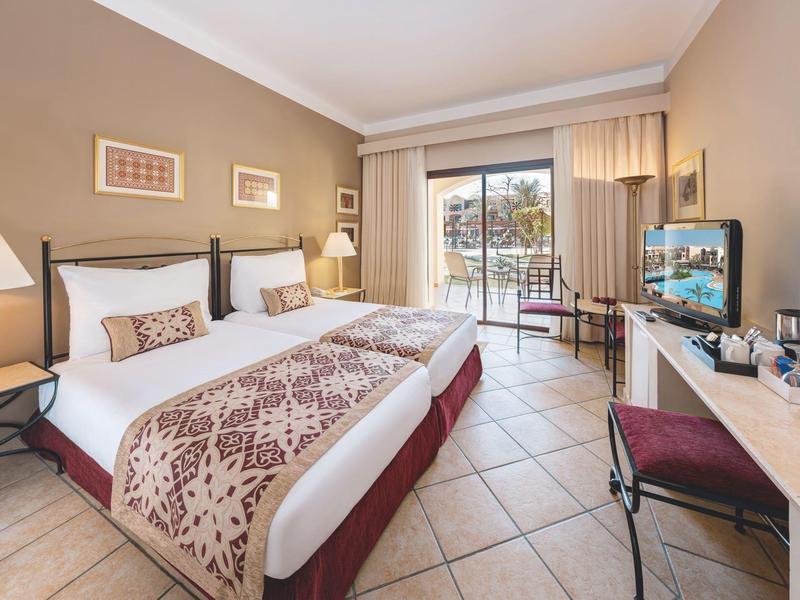 Bright hotel room with double bed, balcony, tiled floor, and modern TV.