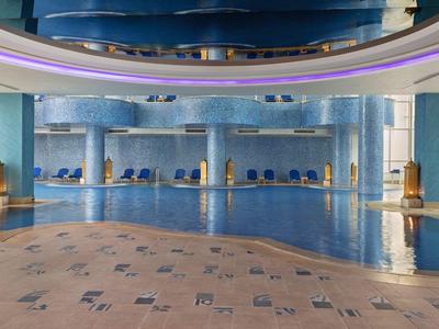 Großes Hallenbad mit blauem Wasser, Säulen und blauen Liegestühlen in hellem, modernem Raum.