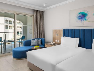 Helles Hotelzimmer mit zwei weißen Betten, blauer Couch, Balkon und moderner Wandkunst.