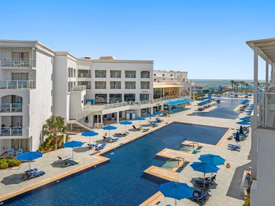 Großer Außenpool mit blauen Liegen und Sonnenschirmen zwischen weißen Hotelgebäuden unter blauem Himmel.