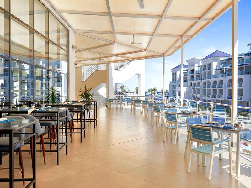 Lichtdurchflutete Terrasse mit Holzterrasse, Glasgeländer und Tischen mit Stühlen.