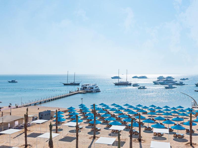 Strand mit blauen Sonnenschirmen, Promenade aus Holz, Meer mit Booten und klarem Himmel im Hintergrund.