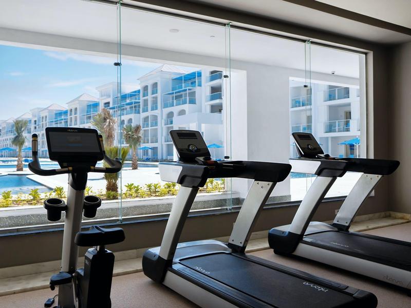 Fitnessraum mit zwei Laufbändern und einem Fahrradergometer vor großen Fenstern mit Blick auf Pool und Gebäude.