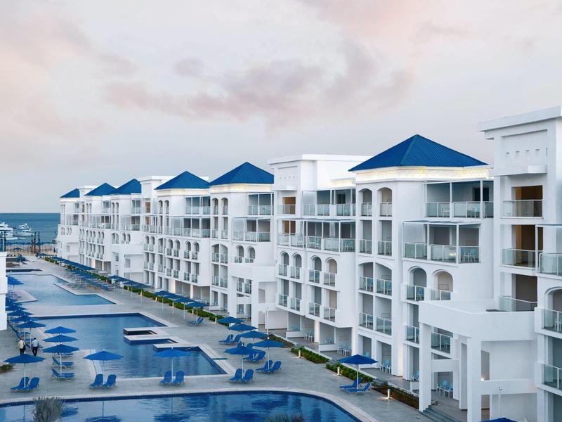 Moderne weiße Apartmenthäuser mit blauen Dächern und Pools unter einem hellen Himmel.