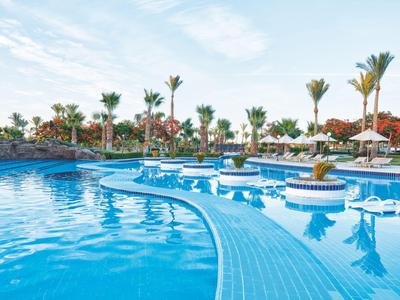 Large hotel pool with palm trees and sun umbrellas under a blue sky.