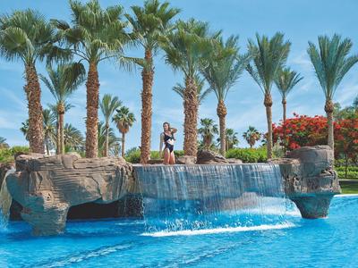 Pool with artificial rock waterfall surrounded by palm trees and colorful vegetation.