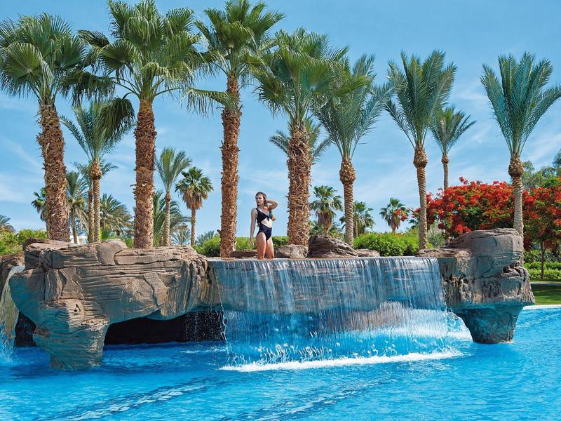 Large pool with waterfall and palm trees under blue sky in tropical setting.