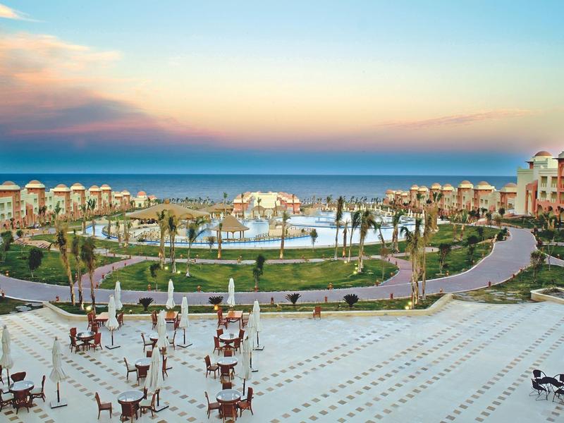 Großer Hotelpool mit vielen Liegestühlen, umgeben von Palmen und Meer im Hintergrund bei Sonnenuntergang.