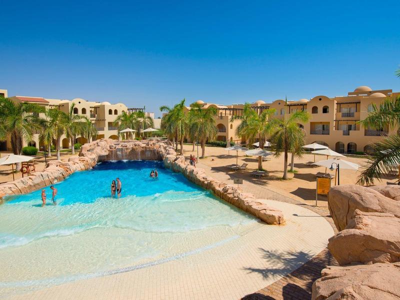 Großer Pool in ruhiger Hotelanlage mit Palmen, Felsen und klar blauem Himmel im Hintergrund.