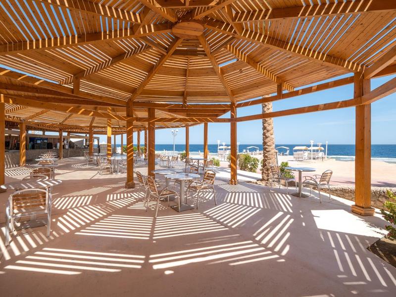 Große Holzkonstruktion mit Schattenspielen, Tischen und Blick auf einen Sandstrand und das Meer.