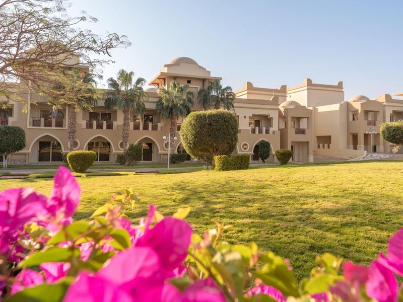 Großes sandfarbenes Gebäude mit Bögen, Palmen und grünem Rasen, vorne pinke Blüten.