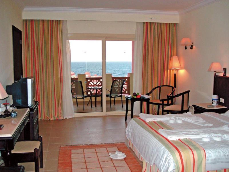 Helles Hotelzimmer mit Doppelbett, zwei Sesseln, Balkon und Meerblick bei Tageslicht.