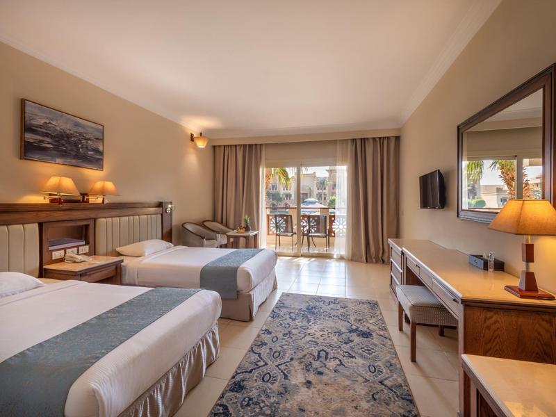 Hotelzimmer mit zwei Einzelbetten, Teppich, Schreibtisch, TV und Balkon mit Blick nach draußen.