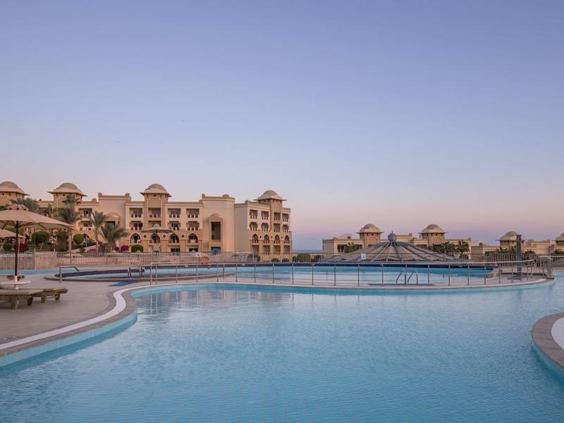 Großer Pool mit klarem Wasser vor einem mehrstöckigen Hotel bei klarem Himmel am Abend.
