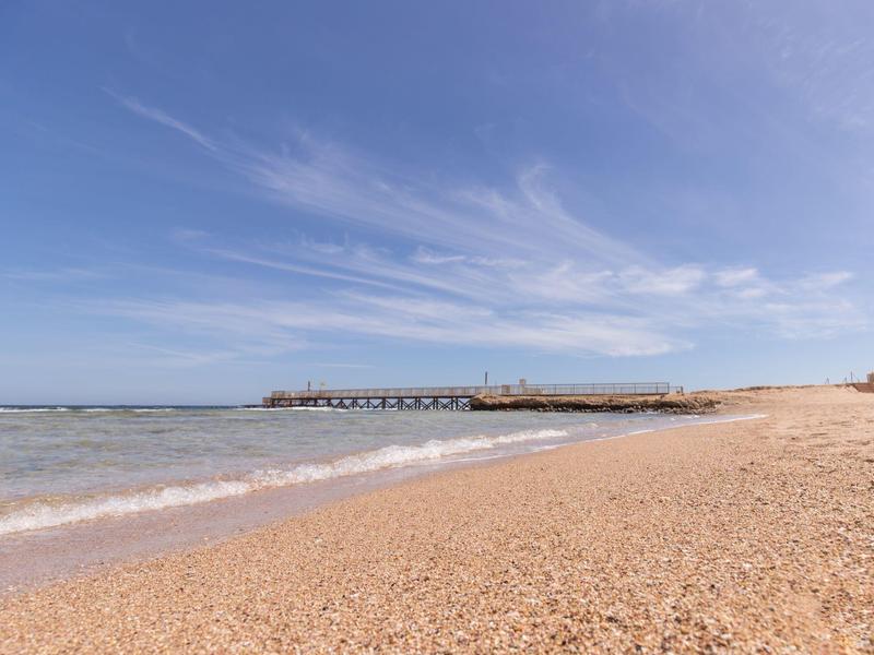 Weicher Sandstrand mit ruhigem Meer und langer Mole unter blauem Himmel mit leichten Wolken.