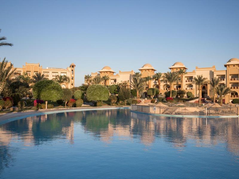 Großer Swimmingpool vor Palmen und beige farbenen Gebäuden unter hellem blauem Himmel.